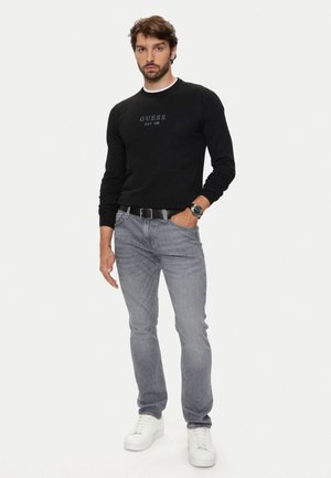 Maglietta nera a maniche lunghe con logo "GUESS EST. 1981", abbinata a jeans grigio chiaro e sneakers bianche, completata da una cintura nera e un orologio.