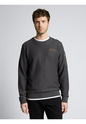 Man die recht vooruit kijkt, draagt een donkergrijs sweatshirt met ronde hals en een klein logo op de borst, en zwarte broek, tegen een lichte effen achtergrond.