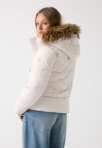 Padded, světle růžová bunda s umělou kožešinou na kapuci, páskem v pase a diagonálním šitím. Model ji nosí s světle modrými džínami a je mírně otočený vzad.