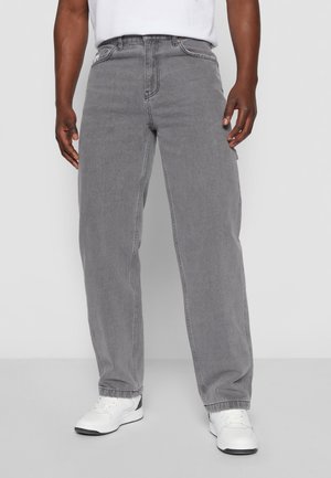 Mann trägt locker sitzende graue Jeans und weiße Turnschuhe, steht vor einem einfarbigen hellgrauen Hintergrund.