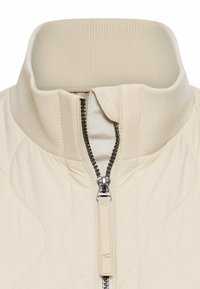 Gestepptes cremefarbenes Jackett mit hohem Kragen, geripptem Hals und einem mittigen Reißverschluss mit metallischen Beschlägen. Glatte Stoffstruktur.