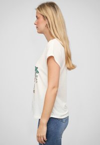 T-shirt en coton blanc à manches courtes, avec un motif graphique coloré sur le devant. Porté avec un jean en denim bleu.