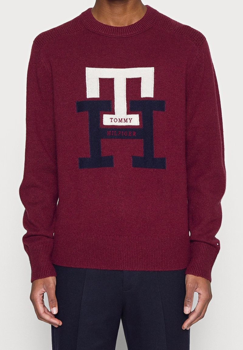 Pull en maille bordeaux avec un grand logo brodé blanc et marine. Encolure ronde et poignets côtelés avec une texture lisse.