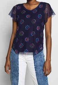Navy blouse with short, sheer sleeves, featuring colorful floral patterns, smooth texture, and slight curved hem. Paired with patterned jeans.