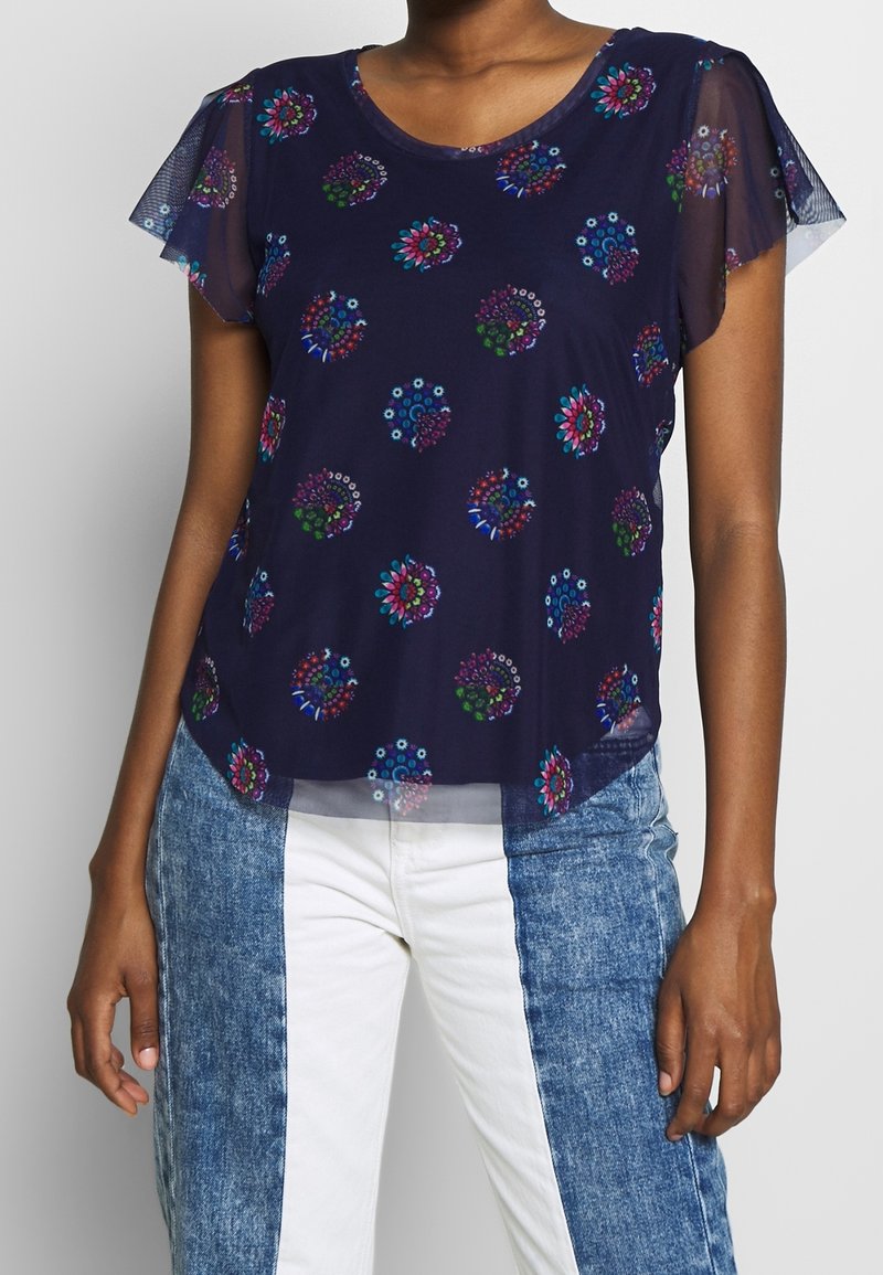 Navy blouse with short, sheer sleeves, featuring colorful floral patterns, smooth texture, and slight curved hem. Paired with patterned jeans.