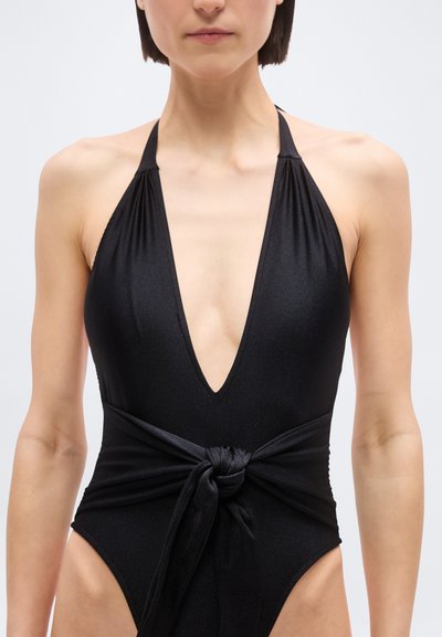 Γυναίκα με μαύρο ολόσωμο μαγιό τύπου halter με βαθύ V και υφασμάτινη ζώνη δεμένη με κόμπο στη μέση, στέκεται μπροστά σε απλό λευκό φόντο.