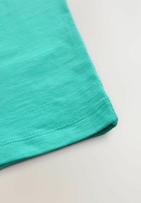 Türkisfarbener Baumwollstoff mit glatter Textur, mit gesäumtem Rand und minimalem Nähedetail, aus einem Winkel betrachtet.