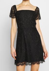 Abito di pizzo nero con scollatura quadrata, maniche corte in pizzo e orlo ondulato. Presenta motivi floreali e una silhouette a vita aderente.