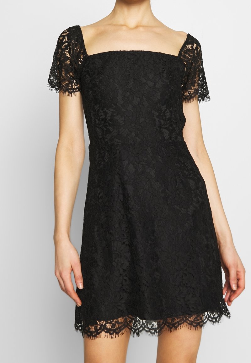 Abito di pizzo nero con scollatura quadrata, maniche corte in pizzo e orlo ondulato. Presenta motivi floreali e una silhouette a vita aderente.