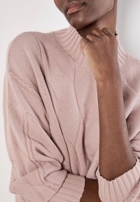 Heller rosa gestrickter Pullover mit hohem Rippenkragen, der ein strukturiertes Muster und kleine silberne Verzierungen aufweist.