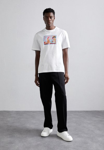 Camiseta de algodón blanca con un estampado gráfico colorido centrado en el pecho, combinada con pantalones negros y zapatillas blancas.