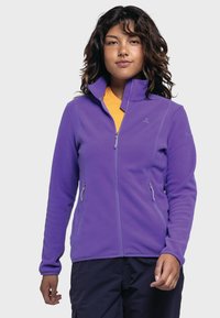 Lila Fleece-Jacke mit hohem Kragen, mit Reißverschlusstaschen an den Seiten und kontrastierenden Nähten. Getragen über einem gelben Shirt mit Logodetails.