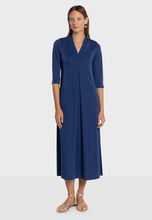 Robe longue bleu marine en tissu doux, avec un décolleté en V, des manches trois-quarts, et un design plissé, associée à des sandales argentées à lanières.