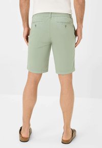 Pantaloni corte verde chiaro con una texture liscia, tasche posteriori con bottoni e un taglio dritto. Indossati con sandali scivolati marroni.