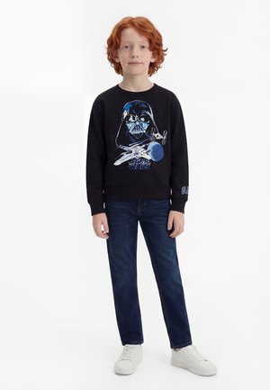 Κόκκινομυαλό παιδί που φοράει μαύρο φούτερ Star Wars, σκούρο μπλε τζιν και λευκά αθλητικά παπούτσια, στέκεται μπροστά σε ένα λευκό φόντο.