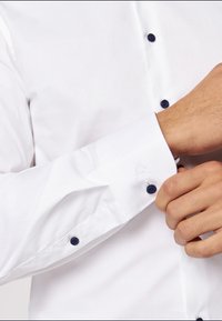 Weißes Hemd mit langen Ärmeln, ausgestattet mit dunkelblauen Knöpfen und Manschetten. Glatte Textur mit einem kleinen Logo auf der Manschette. Taillierte Passform.