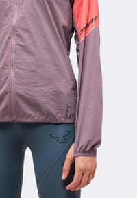Gros plan d'une personne portant une veste zippée violette avec des détails corail et un legging bleu comportant un logo de visage d'animal noir.