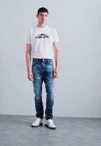 White T-shirt with black logo, blue distressed denim jeans, and white sneakers with black stripes. Simple design, fitted shape.