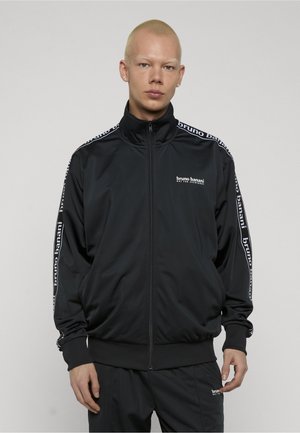 Ung person med kort blondt hår iført en sort Bruno Banani lynlåstrøje med logo på bryst og ærmer.