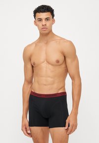 Schwarze Boxer-Briefs mit einem bordeauxroten elastischen Bund, der das Logo "BJÖRN BORG" trägt. Der Stoff wirkt weich und sitzt eng. Schlichte Gestaltung.