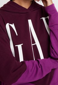 Felpa bordeaux con testo bianco "GAP". Presenta maniche viola, una tasca a marsupio frontale e un tessuto morbido e testurizzato con cappuccio regolabile.