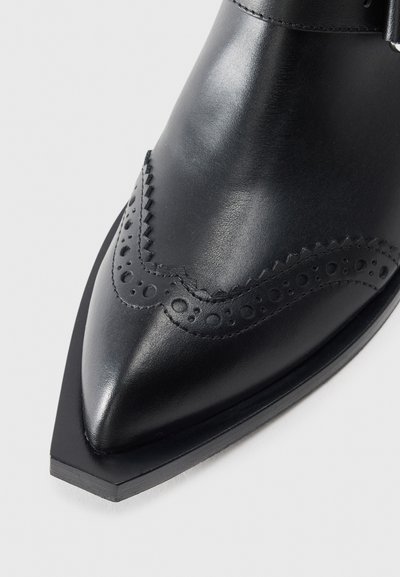 Melns ādas kurpes ar norādītu pirkstu, dekoratīvo caurumošanas brogu apdari un zemu kaudzes papēdi uz baltas fona.