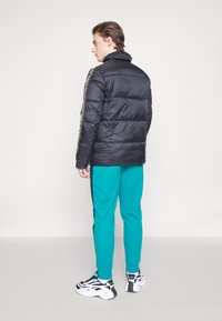 Sort puff jakke med høj krave og horisontal quiltet design, sammen med teal træningsbukser med sorte sidelinjer. Hvide fodtøj.