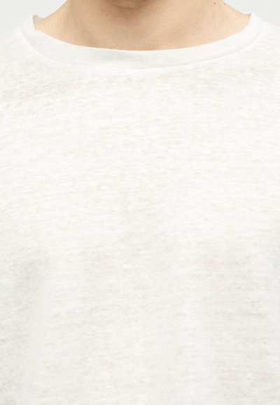 Κοντινό πλάνο στον λαιμό και το πάνω μέρος του στήθους ενός ατόμου που φοράει μια απλή λευκή μπλούζα με στρογγυλή λαιμόκοψη και απαλό υφασμάτινο υλικό.