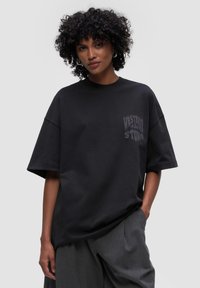 Czarna, oversize koszulka z miękkiego materiału, z okrągłym dekoltem i graficznym nadrukiem po lewej stronie. Połączona z jasnoszarymi spodniami.