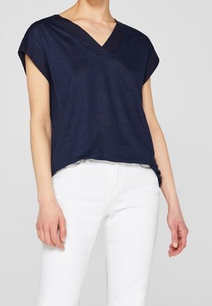 Mujer que lleva una camiseta azul oscuro de manga corta con cuello en V, metida ligeramente dentro de pantalones blancos, de pie con los brazos relajados a los lados.