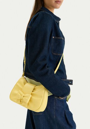 Kobieta ubrana w ciemnoniebieską kurtkę i spodnie dżinsowe, niosąca małą żółtą torebkę na ramię z ozdobą w formie kokardy.