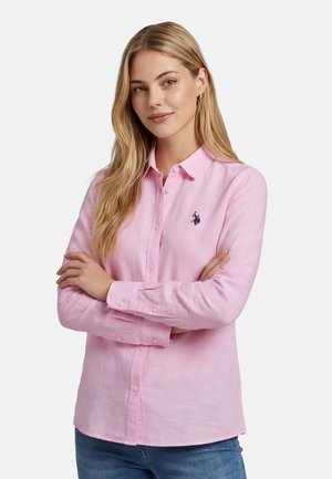 Woman with long blonde hair, wearing a light pink button-up shirt with a small dark logo, arms crossed, and blue jeans against a white background.