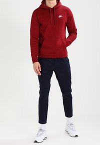 Burgundowa bluza Nike z kieszenią z przodu i białym logo, zestawiona z granatowymi spodniami oraz białymi trampkami z szarymi akcentami i fakturowanymi sznurówkami.