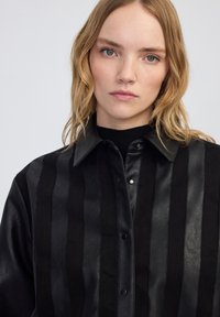 Czarna koszula zrobiona ze skóry i zamszu, charakteryzująca się pionowymi paskami, kołnierzykiem i zatrzaskami, o gładkiej fakturze i błyszczące wykończeniu.