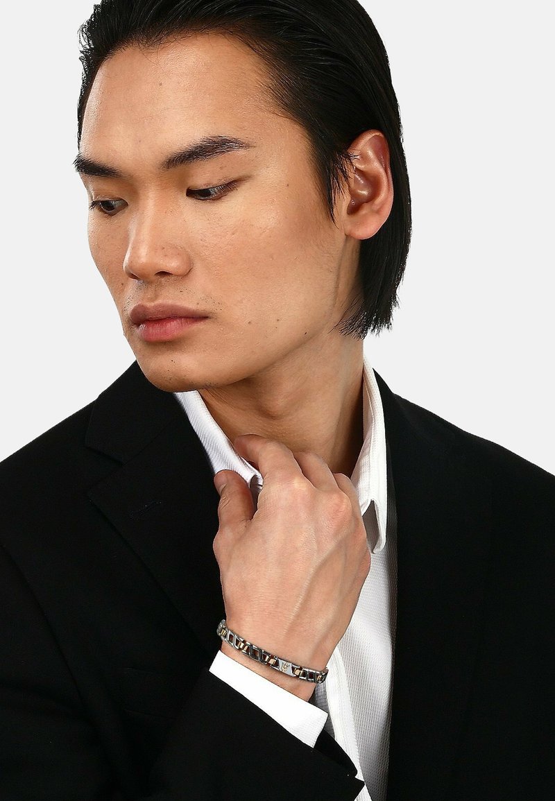Silberfarbener Armreif mit geometrischen Metallakzenten, getragen an einer Hand, die einen Kragen berührt, vor einem schwarzen Anzug mit einem weißen Hemd.