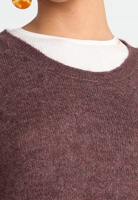 Maglione lavorato a maglia color marrone scuro con colletto rotondo, indossato sopra una camicia bianca a costine. Morbido al tatto con una vestibilità leggermente ampia.