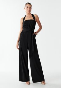 Woman standing wearing a black halter-neck wide-leg jumpsuit with a tied waist belt and gold open-toe heels against a white background.