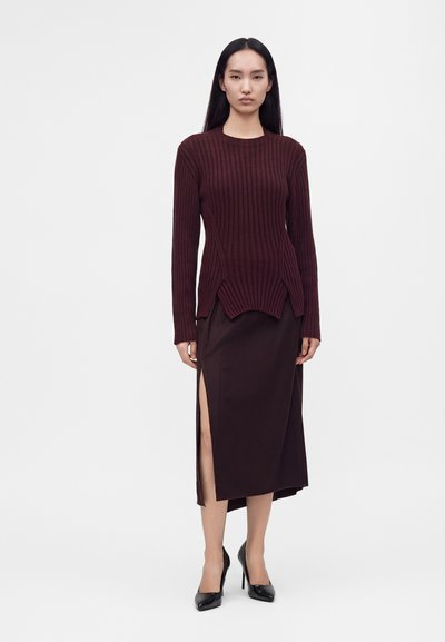 Γυναίκα με σκούρο κόκκινο ριμπ πλεκτό πουλόβερ με μακριά μανίκια και σκούρα φούστα με ψηλή σχισμή, σε συνδυασμό με μαύρα ψηλοτάκουνα παπούτσια.