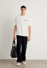 Balta kokvilnas T-kreklu ar "Calvin Klein" logo, saskaņota ar tumši zilibām platām biksēm, bēšām kurpēm un melnu rokassomu. Minimālisma dizains.