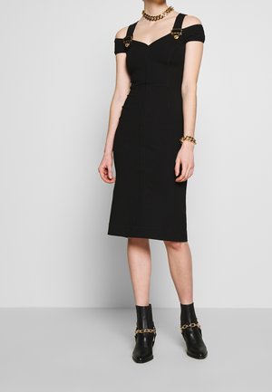 Femme portant une robe noire près du corps à épaules dénudées avec des bretelles à boucle dorée, des bottines noires avec chaînes dorées et des bijoux dorés.