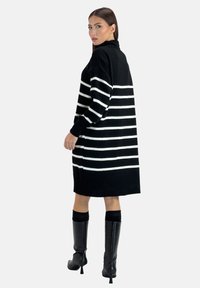 Robe en maille noire à rayures horizontales blanches, manches longues, col montant, ourlet côtelé et fentes latérales ; portée avec des bottes noires montantes jusqu'aux genoux.