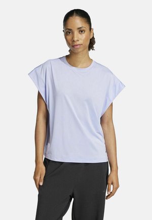 Frau mit zurückgebundenem Haar, die ein lockeres, helllila ärmelloses Oberteil und schwarze Hosen trägt, steht vor einem schlichten weißen Hintergrund.