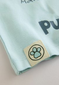 Lichtblauwe katoenen t-shirt met zwarte tekst en een vierkante label met een pootafdruklogo. Zorgvuldige textuur, opgevouwen zoom en subtiele stiksels.