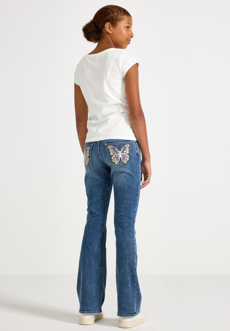 Κορίτσι με λευκό t-shirt και μπλε τζιν με πολύχρωμα σχέδια πεταλούδων στις πίσω τσέπες, στέκεται μπροστά σε απλό φόντο.