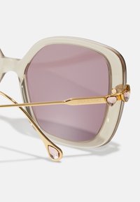 Gafas de sol con montura dorada y acabado beige transparente, con lentes morados. Presentan acentos de cristales Swarovski en forma de lágrima en las patillas.