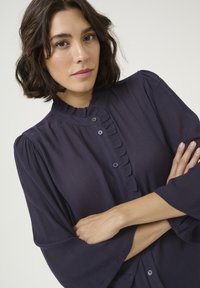 Blouse bleu marine avec col à volants, fermeture à boutons et manches longues et larges. Le tissu texturé ajoute un intérêt visuel.