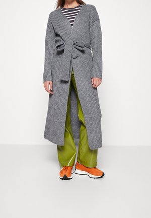 Kvinde iført en lang grå ribstrikket cardigan, bundet ved taljen, grønne bukser med vide ben, stribet top og orange sneakers, stående mod en hvid baggrund.