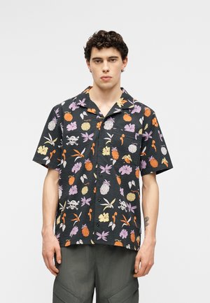 Jonge man die een zwart kort mouw knoopoverhemd draagt met kleurrijke tropische fruit- en bloemenprints, staande tegen een effen lichte achtergrond.