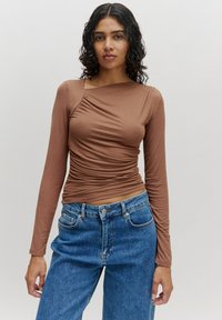 Langärmliges braunes Oberteil mit geraffter Textur an der Vorderseite, ausgestattet mit einem weiten Ausschnitt, kombiniert mit hellblauen, hochgeschnittenen Jeans.