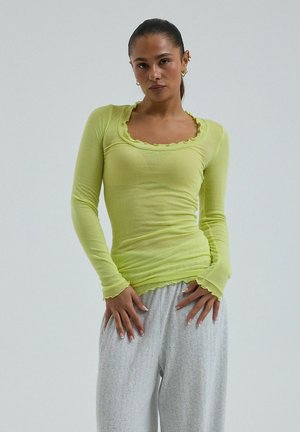 Femme aux cheveux tirés en arrière, portant un haut moulant à manches longues vert citron et un pantalon ample gris clair à motifs, debout avec les mains près des hanches.